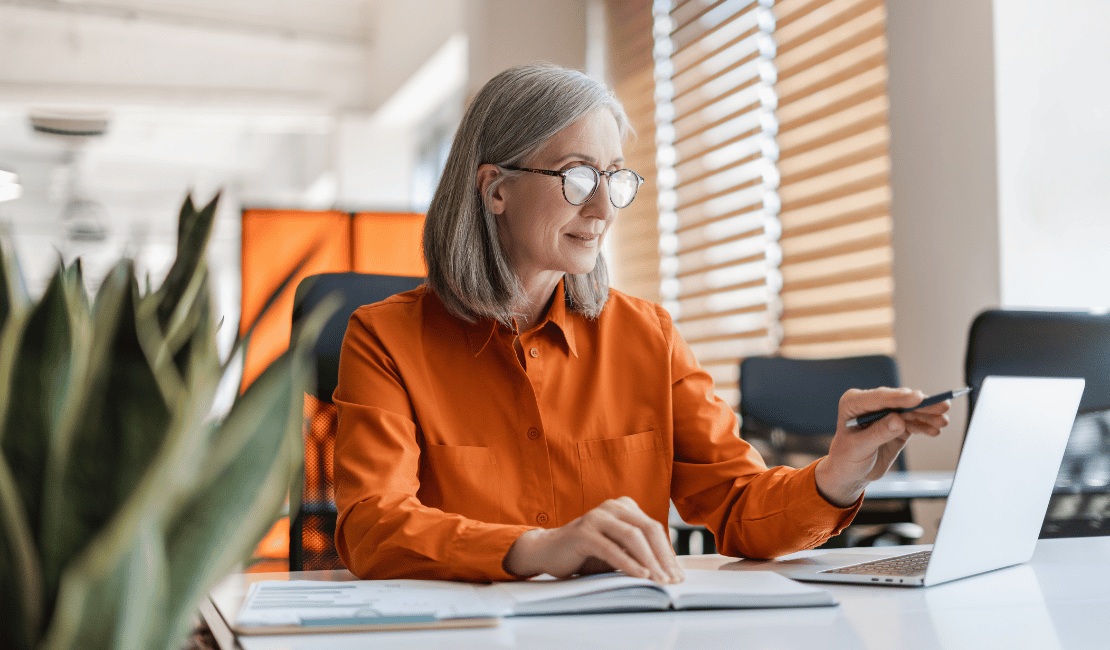 Senior woman using laptop and smartphone in modern office environment.
